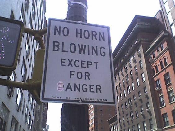 This meme features a photo of a traffic sign that says "No horn blowing except for anger" with a red "No" symbol in a circle. The sign is displayed in a city setting, likely in a busy area with many people and vehicles. The sign's unusual message and use of a red "No" symbol, typically used to indicate a rule or warning, are likely meant to be humorous and satirical.